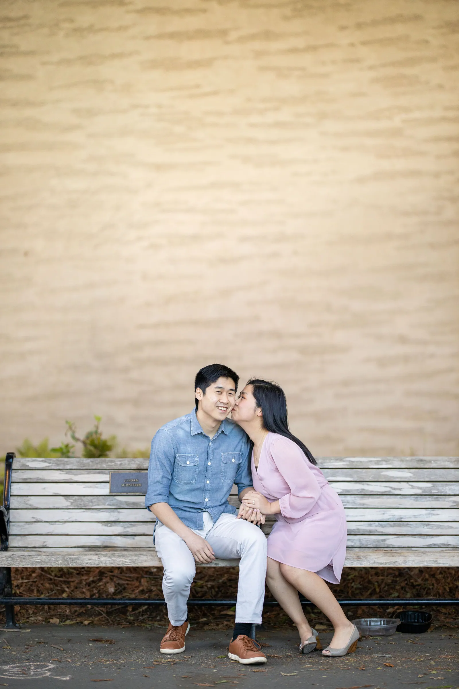 Couple on a wooden bench in front of a cream wall, her kissing his cheek