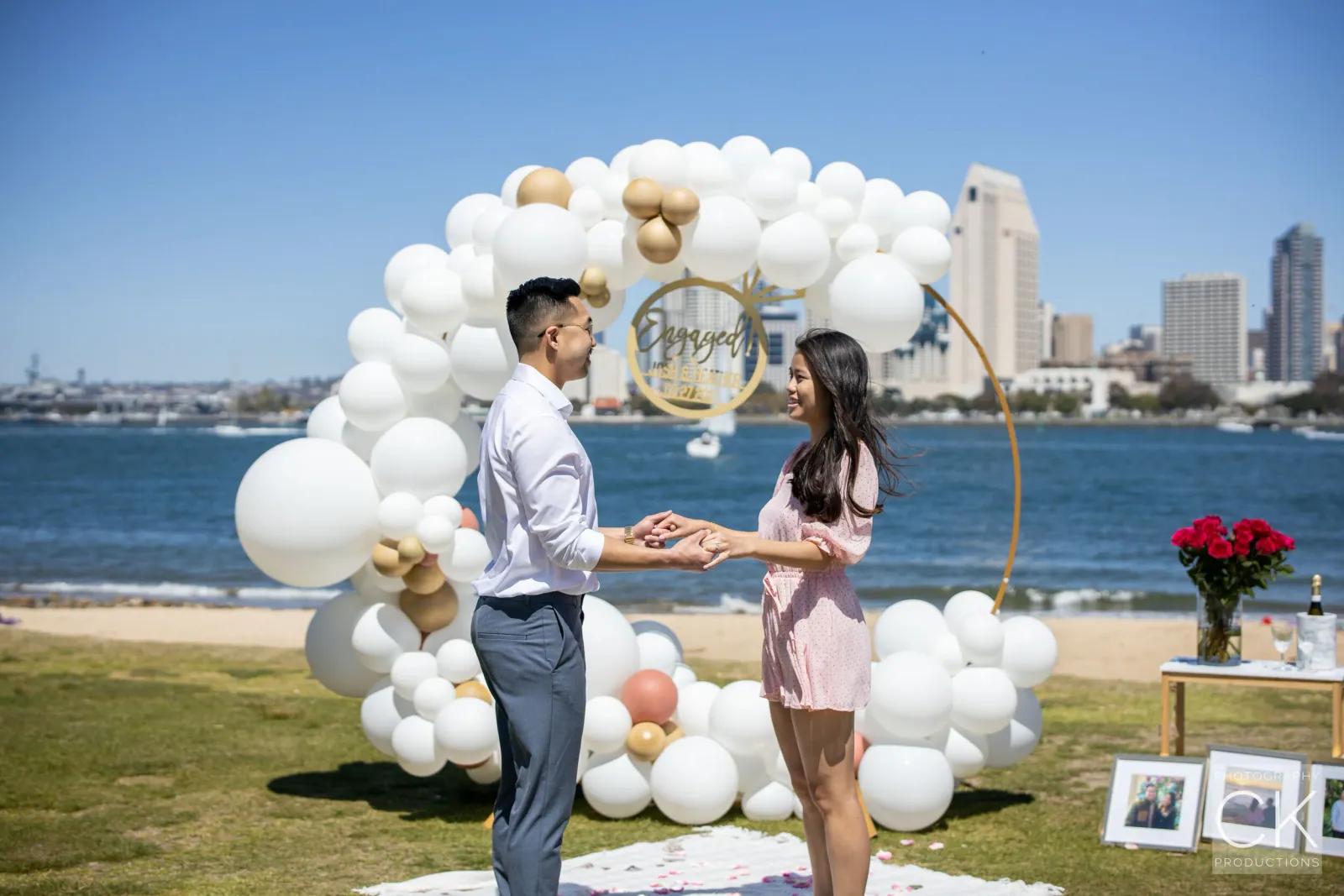 Proposal by a white and gold balloon arch on a waterfront lawn with the city skyline behind