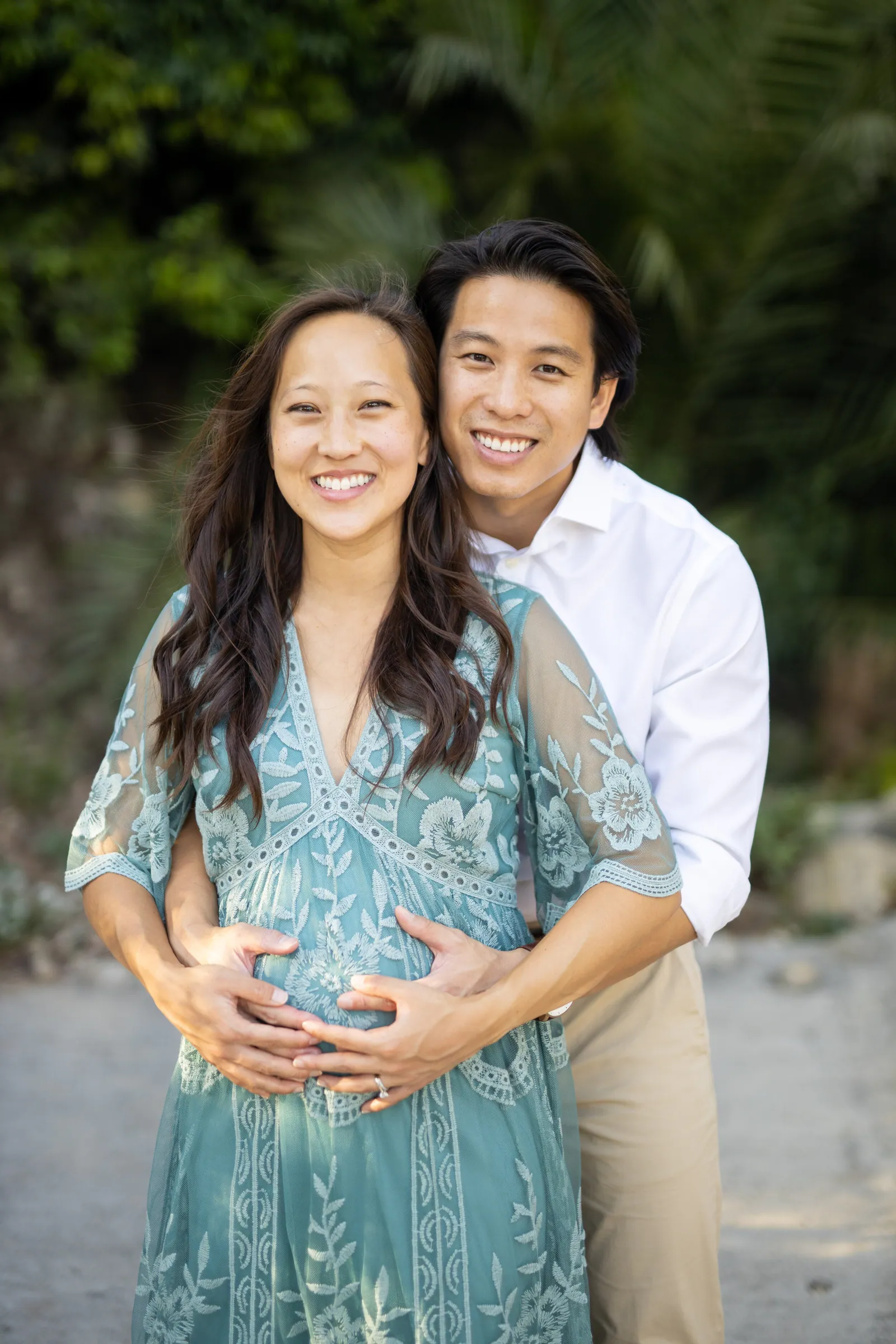 Expectant couple at the coast, her in a teal lace dress, his hands on her belly