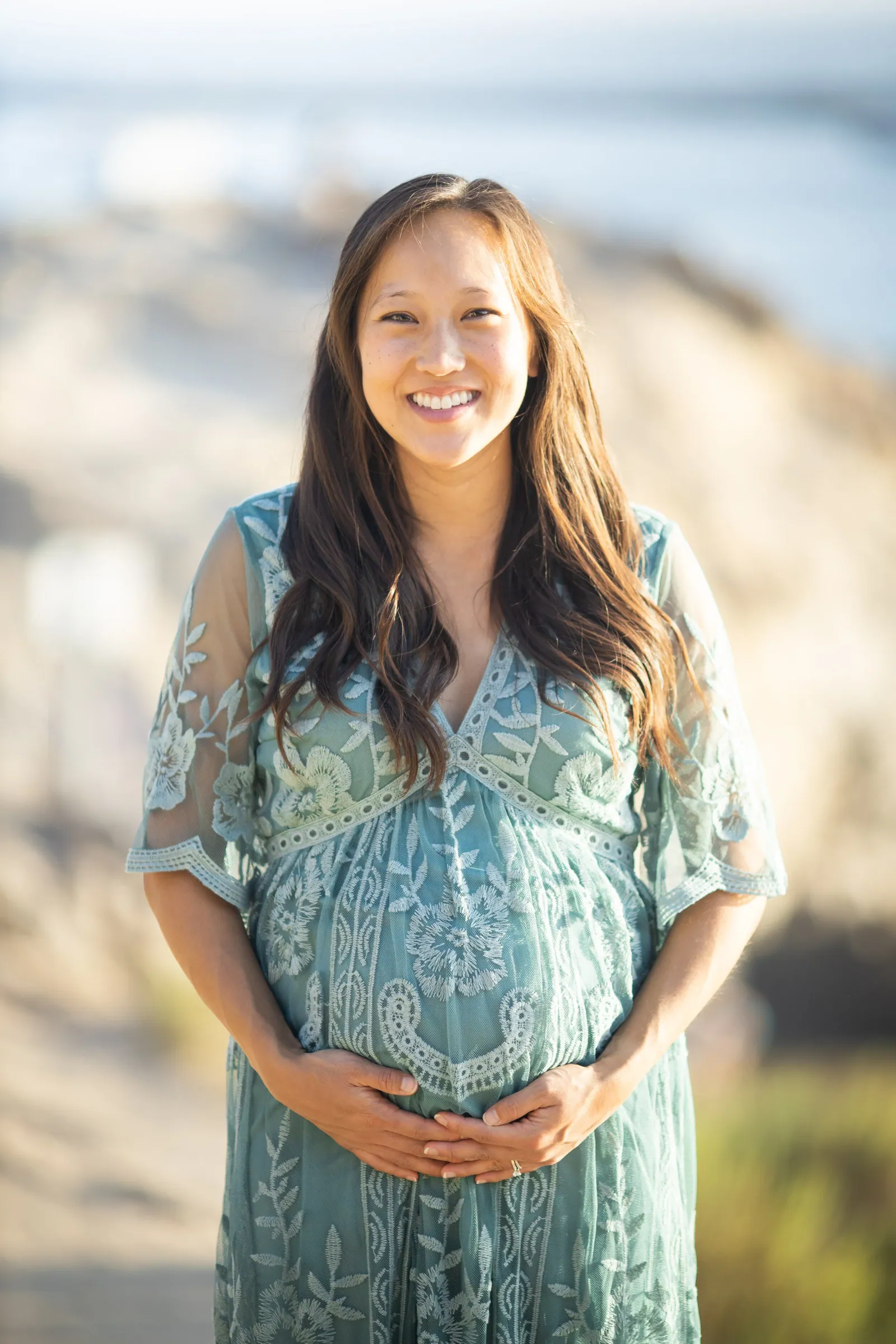 Smiling pregnant woman in a teal lace dress, hands on her belly, with a blurred coast behind