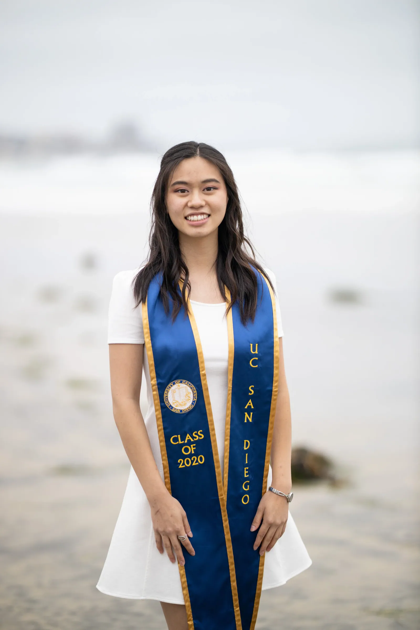 Graduate in a white dress and blue stole standing on a misty beach