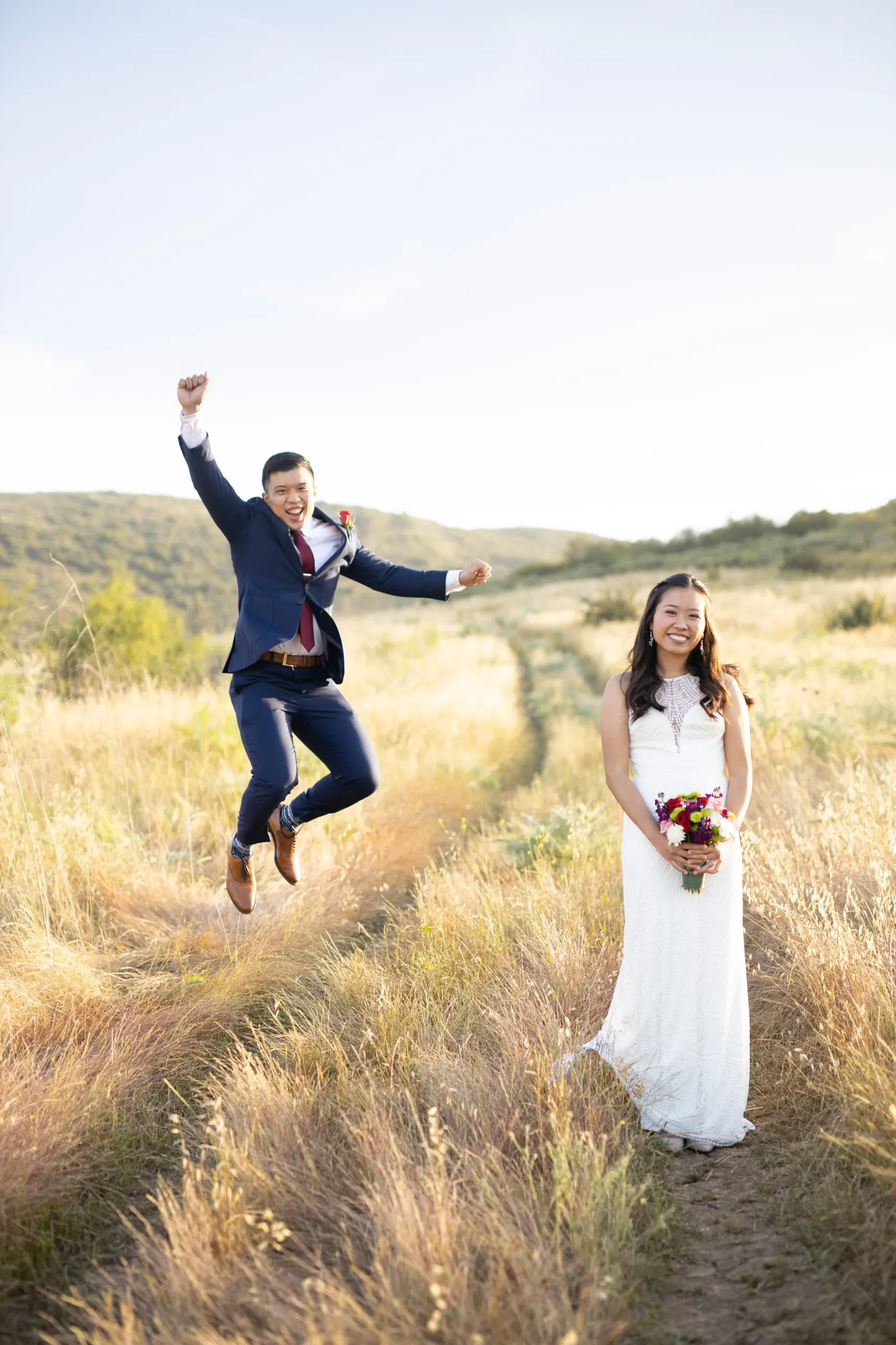 Groom leaping in the air on a grassy trail while the bride stands holding her bouquet
