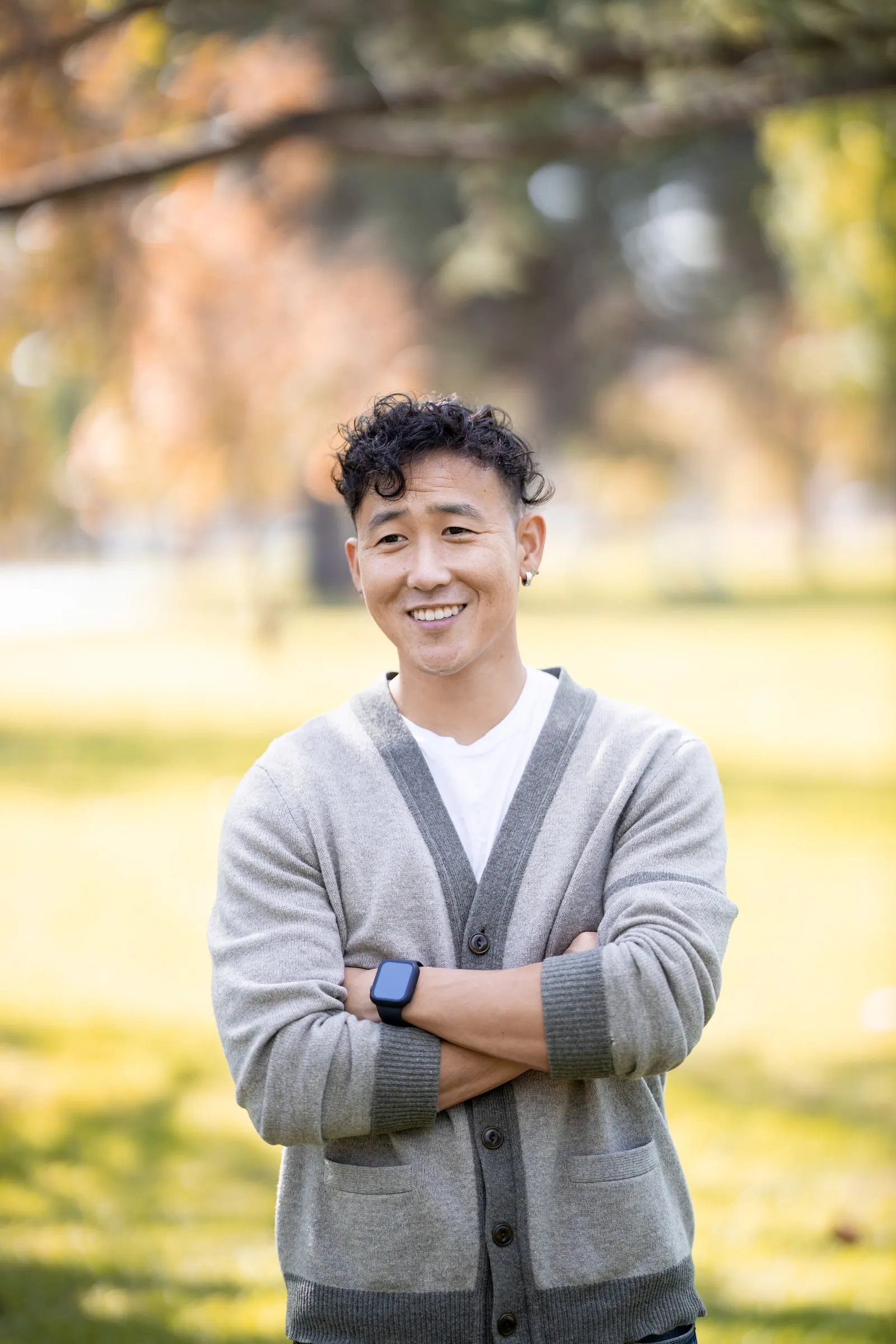 Man in a grey cardigan smiling with arms crossed in a park