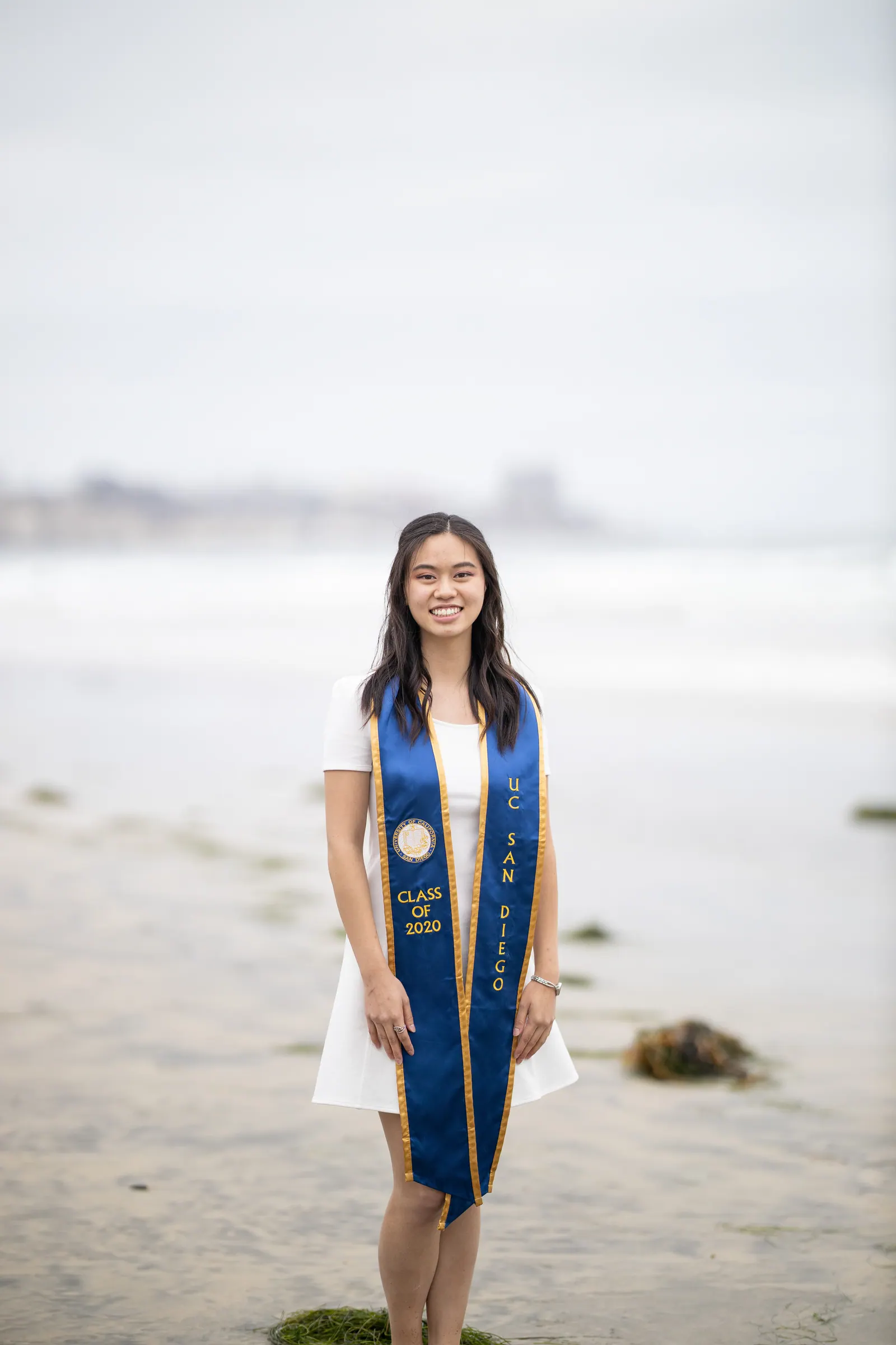 Graduate in a white dress and blue stole on misty sand