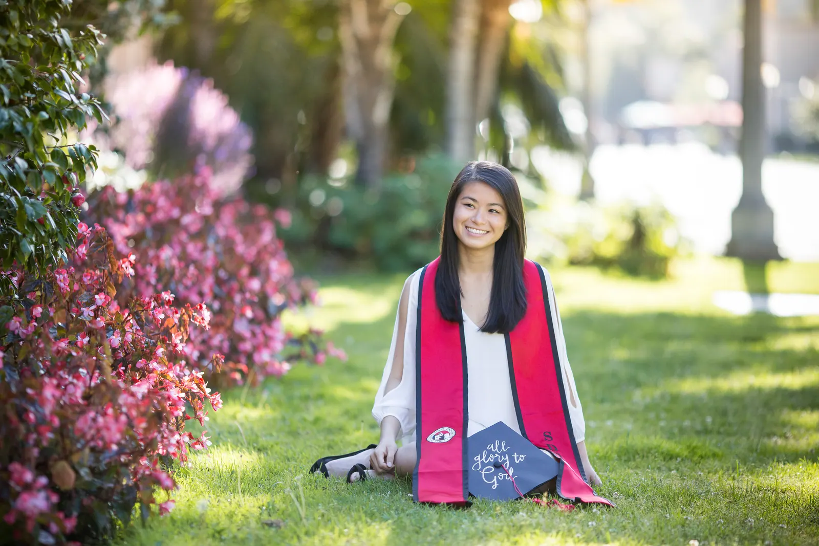 Graduate in a red stole sitting on grass among flowers