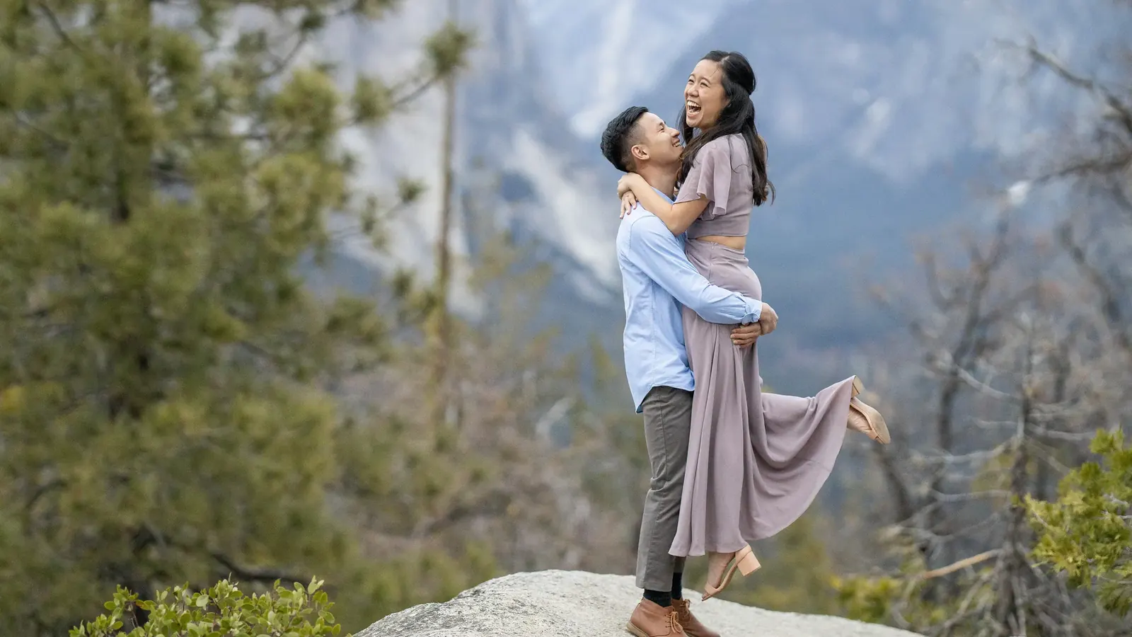 A couple embracing on a granite outcrop above Yosemite Valley: he lifts her off the ground as she laughs, with pine trees and snowy mountains behind them
