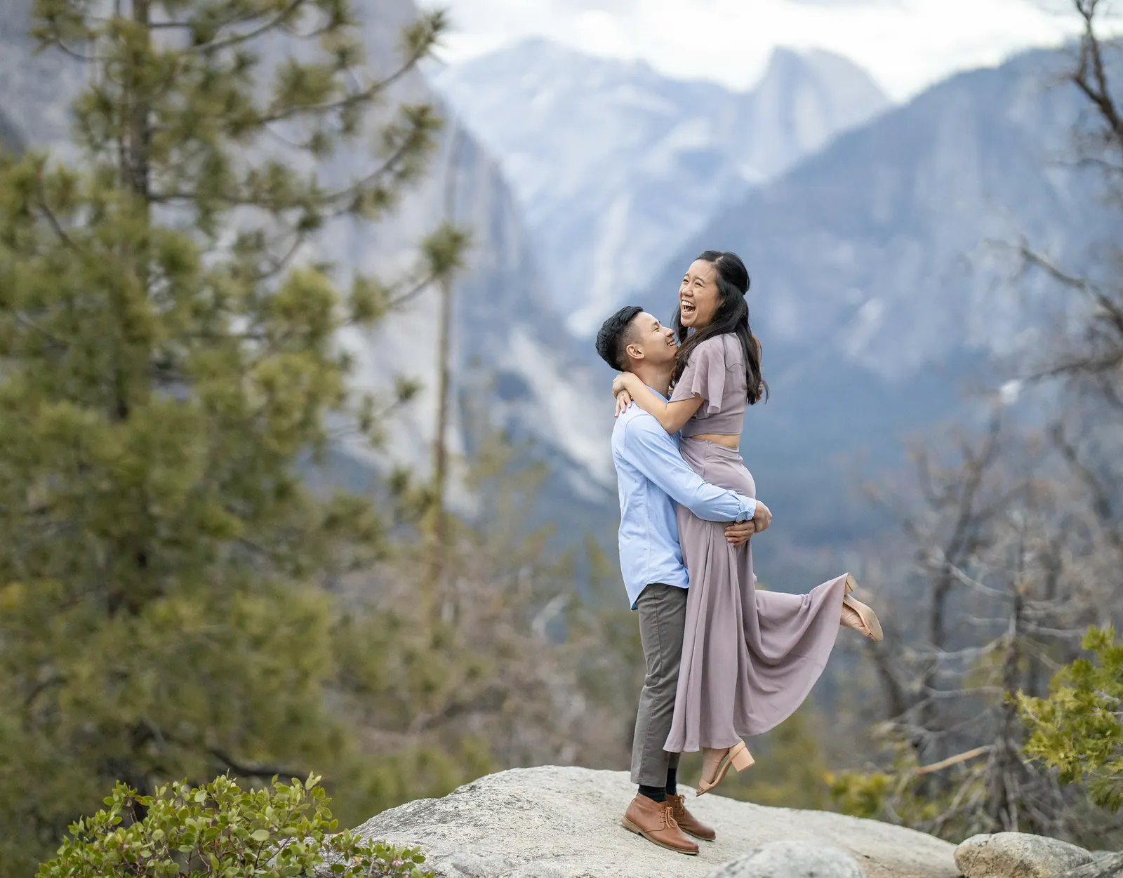 A couple laughing on a granite outcrop above Yosemite Valley as he lifts her off the ground, with pines and snowy peaks behind them