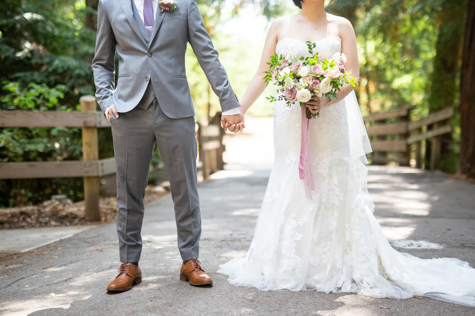 Groom and bride holding hands on a forest path, bouquet in hand